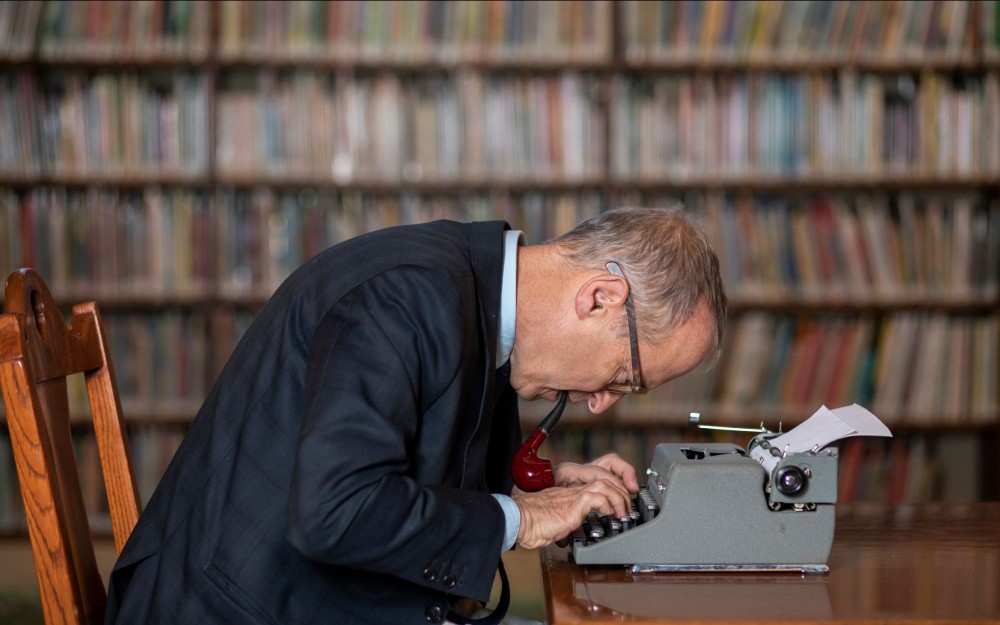 Man in suit hunched over a typewriter with a pipe in his mouth at a library table, bookshelves blurred behind