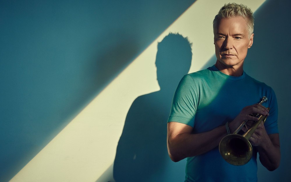 Portrait of an older man holding a trumpet, teal shirt, dramatic shadow.