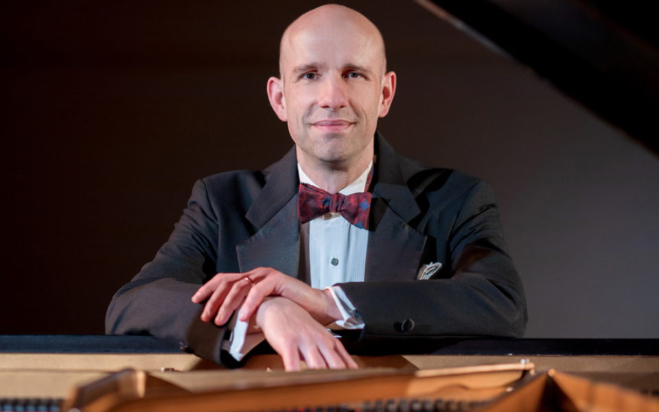 Christopher Taylor in a tuxedo leans on a grand piano with his hands crossed.