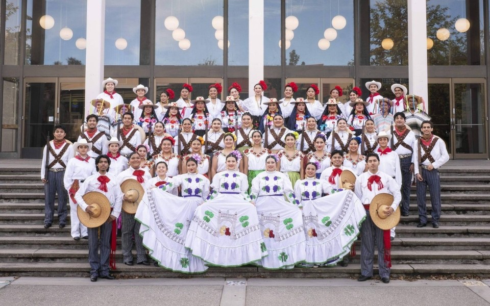 A large group of performers in traditional clothing poses on steps, with vibrant dresses and sombreros.