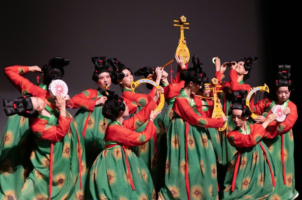 A group of dancers in vibrant green and red costumes perform on stage, holding traditional props.