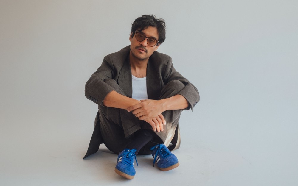 Young Man in blazer and sunglasses sitting cross-legged on plain studio floor, blue sneakers.