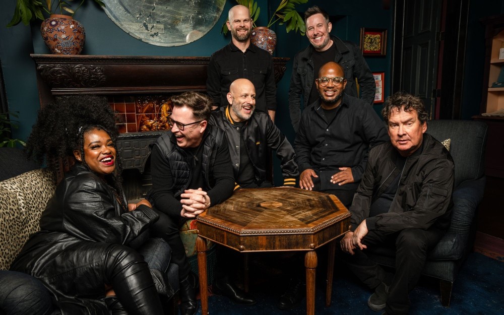Group portrait of seven people gathered around a small wooden table in a dark cozy room