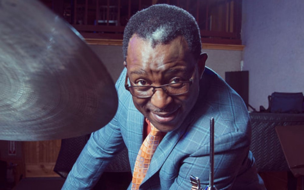 Herlin Riley in a blue suit and an orange tie leans by a drum cymbal