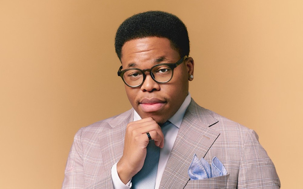 Isaiah J. Thompson smirking at the camera in a tan suit and glasses, hand on chin, in front of a beige background