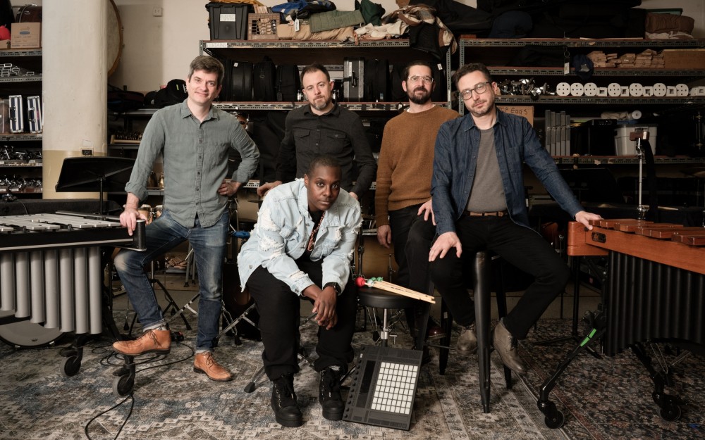 Five musicians pose in a music studio with various instruments and equipment in the background.