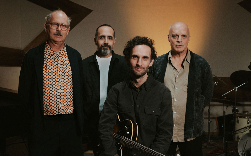 Julian Lage Quartet with John Medeski, Jorge Roeder, & Kenny Wollesen standing behind Julian Lage who is sitting and holding an electric guitar.