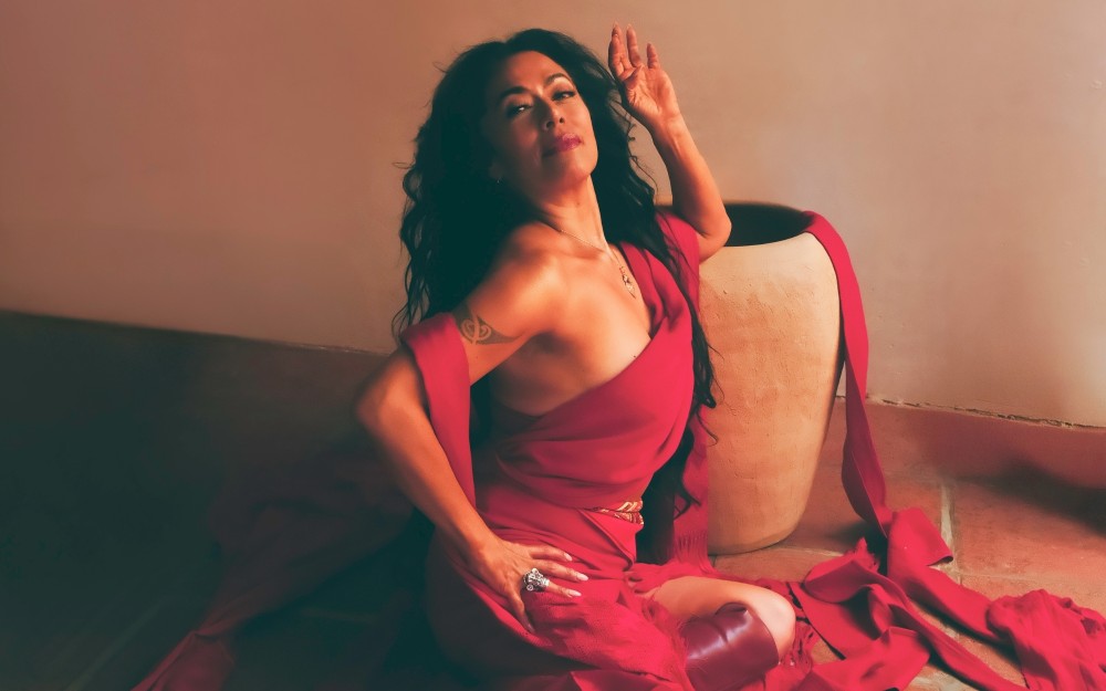 Lila Downs wears a flowing red outfit and poses near a large clay pot.