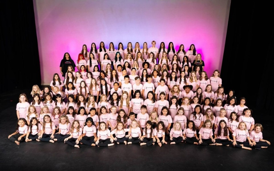 A large group of girls in matching outfits posing together on stage with a pink backdrop.