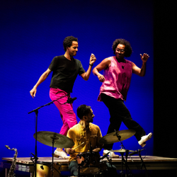Two dancers tapping while drummer plays against blue backdrop