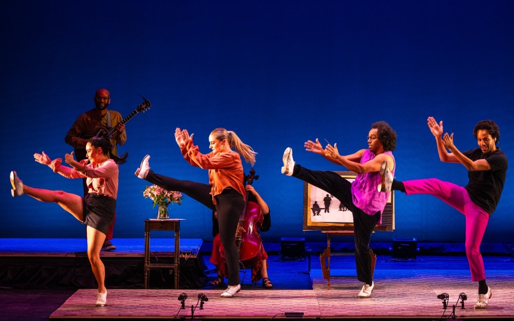 Four dancers mid-kick on blue-lit stage in colorful outfits, with a guitarist and cellist behind.