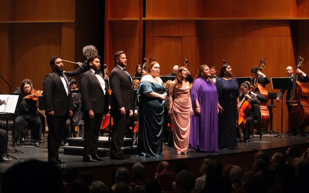A group of six vocal performers on stage, dressed elegantly, with an orchestra in the background.