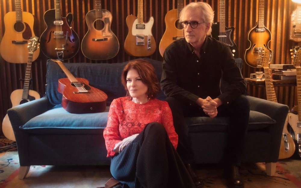 Rosanne Cash sits in front of John Leventhal on blue couch in guitar-filled studio