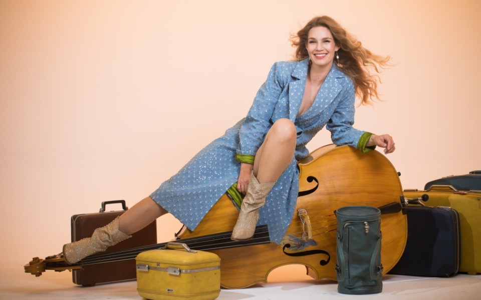 Shelby Means in a blue polka-dot long coat poses on a double bass surrounded by luggage.