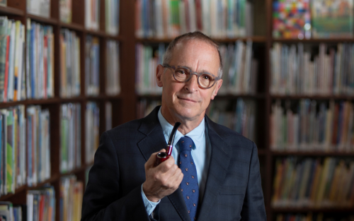 David Sedaris in suit and glasses holding a pipe in a library.