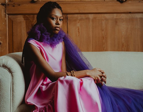 Woman in pink satin gown with long purple tulle drape, seated on sofa, contemplative