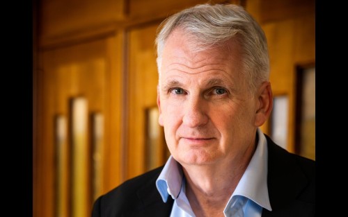 Gray-haired man in dark blazer smiling slightly in front of wooden doors