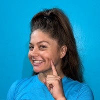 Smiling young woman pointing to her cheek, with a ponytail against a bright blue background