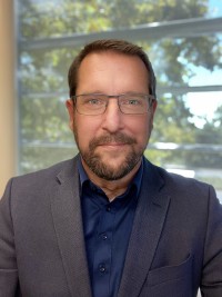Jeremy Ganter in glasses and navy blazer; headshot photograph with window and trees behind