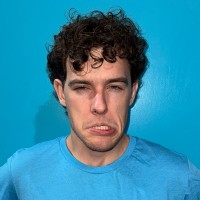 Man with curly hair making an exaggerated grimace against a bright blue wall