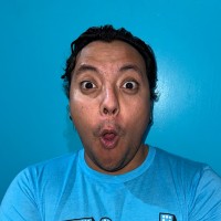 Surprised man with short dark hair, wide eyes, and an open mouth, against a bright blue background.