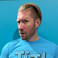 Man with slicked-back blond hair, stubble and a surprised look on a bright blue background