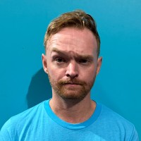 Light-skinned man with short red hair and a quizzical expression on a bright blue background