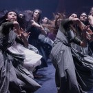 Dancers in flowing gray and white dresses in dynamic contemporary performance on dim stage