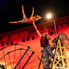 Man balances woman with one arm high above on red-lit circus stage.
