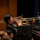 Trombonists in black performing with brass trombones on a wood-paneled stage