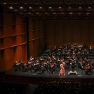 Orchestra with conductor and soloist in pink on a wood-paneled concert stage