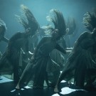 Multiple dancers in flowing gowns, hair and fabric mid-swish under moody blue-green stage lighting
