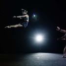 A dancer leaps in mid-air while other performers reach out toward them in a spotlight.