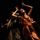 Lead dancer in orange with arms raised over head as if placing a crown, amid shadowed ensemble on dark stage