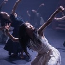 Central dancer in white with face paint reaching upward amid dark-clad ensemble