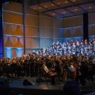 Alumni and University Choruses performs on stage with the UC Davis Symphony Orchestra, illuminated by soft blue and warm lights.