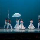 Six dancers in white costumes perform on stage with props including a parasol and poles.