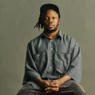 Portrait of Ambrose Akinmusire wearing a lose grey shirt seated against a neutral background.