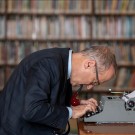 Man in suit hunched over a typewriter with a pipe in his mouth at a library table, bookshelves blurred behind