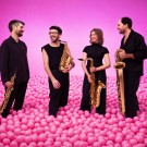 Four saxophonists in black standing in a sea of pink balls against pink backdrop