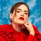 Camila Fernández posed with hand on chin, red ruffled blouse, and a blue backdrop.