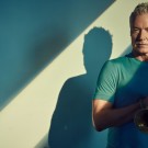 Portrait of an older man holding a trumpet, teal shirt, dramatic shadow.