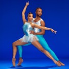 Two ballet dancers in teal costumes; male supports female against solid blue studio backdrop