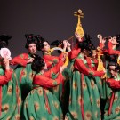 A group of dancers in vibrant green and red costumes perform on stage, holding traditional props.