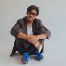 Young Man in blazer and sunglasses sitting cross-legged on plain studio floor, blue sneakers.