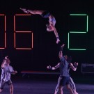 A performer is being tossed in mid-air by a group of dancers on stage, with large digital numbers in the background.