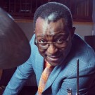 Herlin Riley in a blue suit and an orange tie leans by a drum cymbal