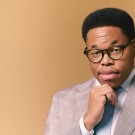 Isaiah J. Thompson smirking at the camera in a tan suit and glasses, hand on chin, in front of a beige background