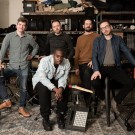 Five musicians pose in a music studio with various instruments and equipment in the background.
