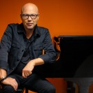 John Beasley in glasses leaning on black grand piano, orange background